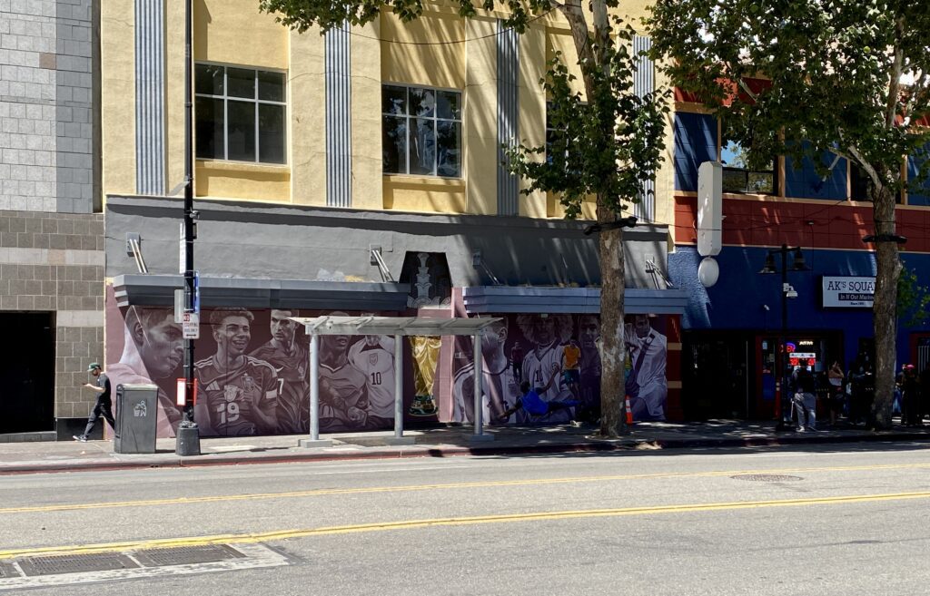 Photo of a mural beside the street showing nine soccer stars and the FIFA world cup trophy next to a repainted blue and red building facade.