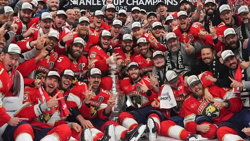 A jubilant hockey team in red jerseys celebrates their Stanley Cup win, with Evan Rodrigues holding the trophy aloft.