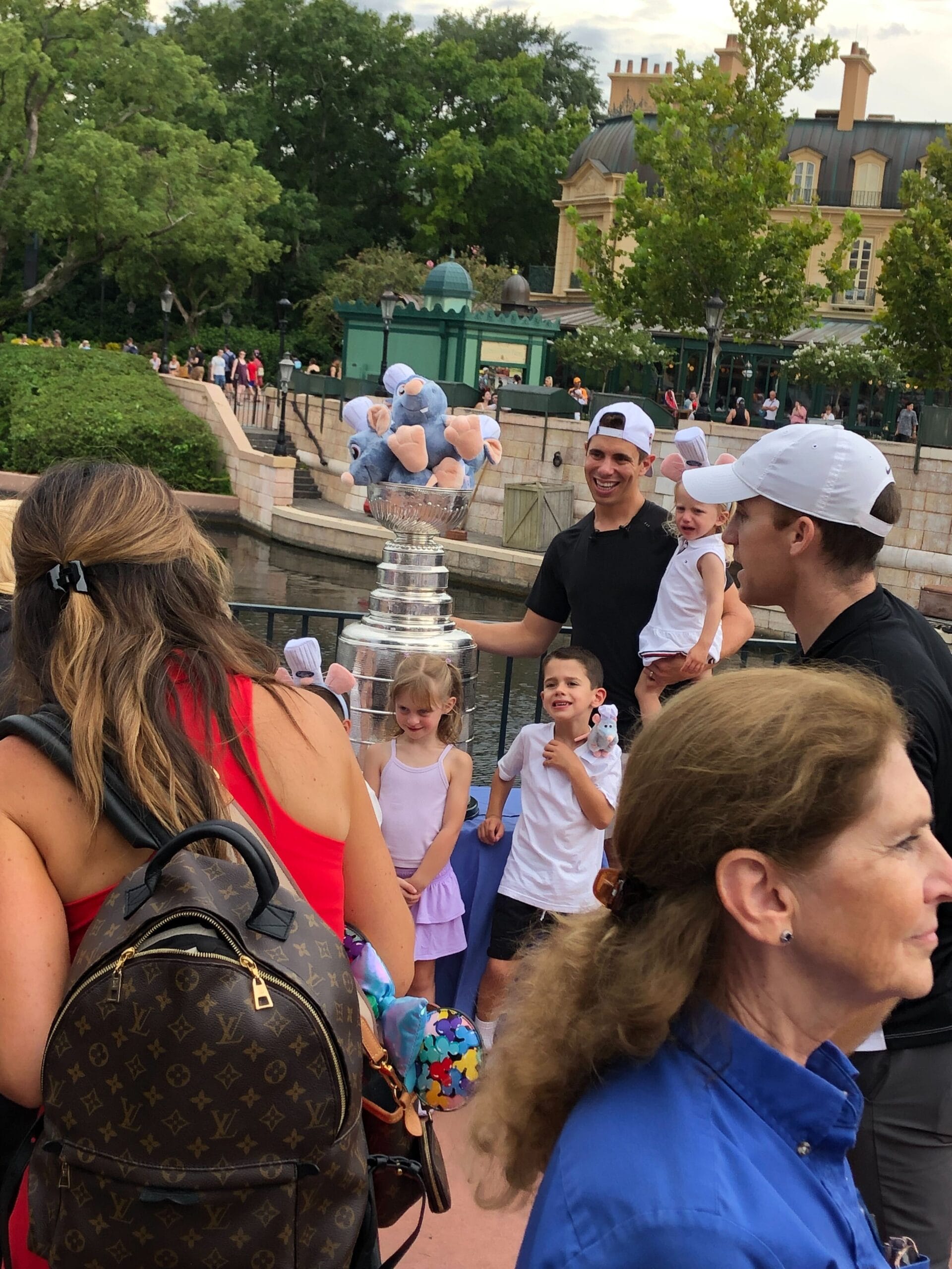Evan Rodrigues holds a large trophy, surrounded by smiling children and adults outdoors in a park area.