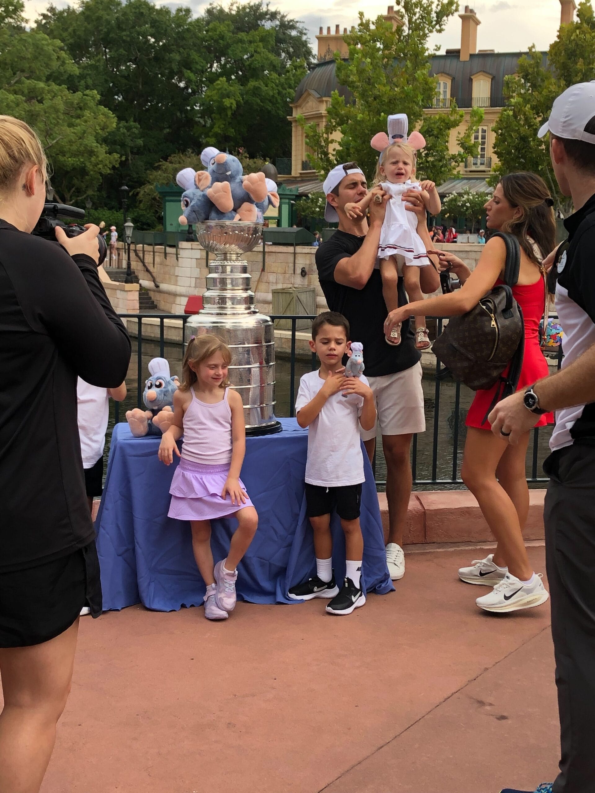 A family with young children poses by the Stanley Cup outdoors, surrounded by blue plush toys.