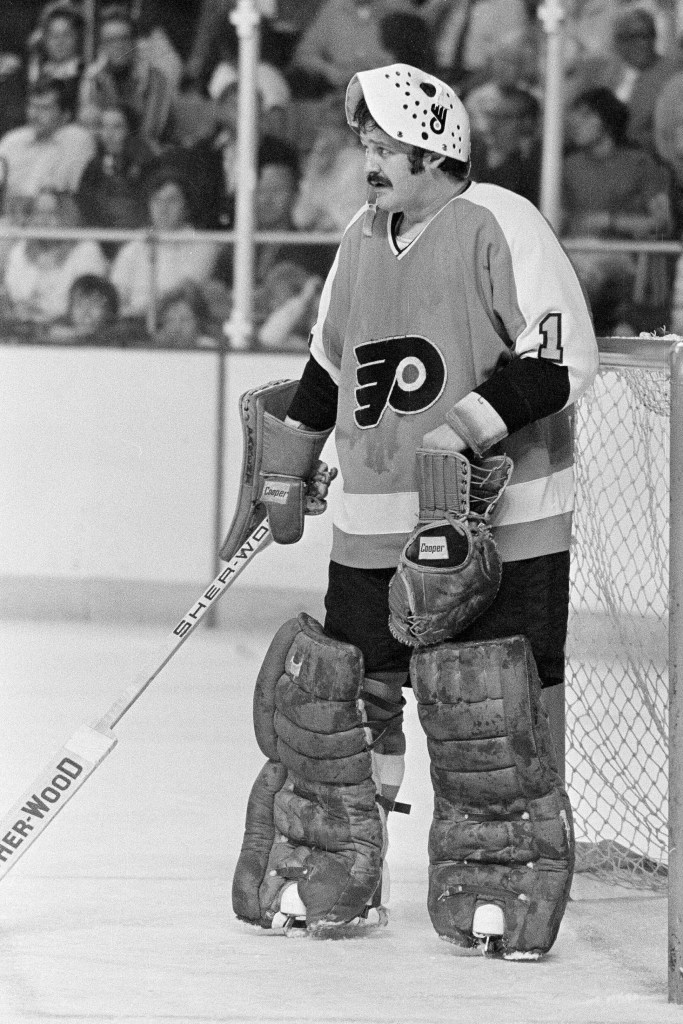 Philadelphia Flyers goalie Bernie Parent wearing his mask pulled up, holding a stick and a catching glove, during a game.