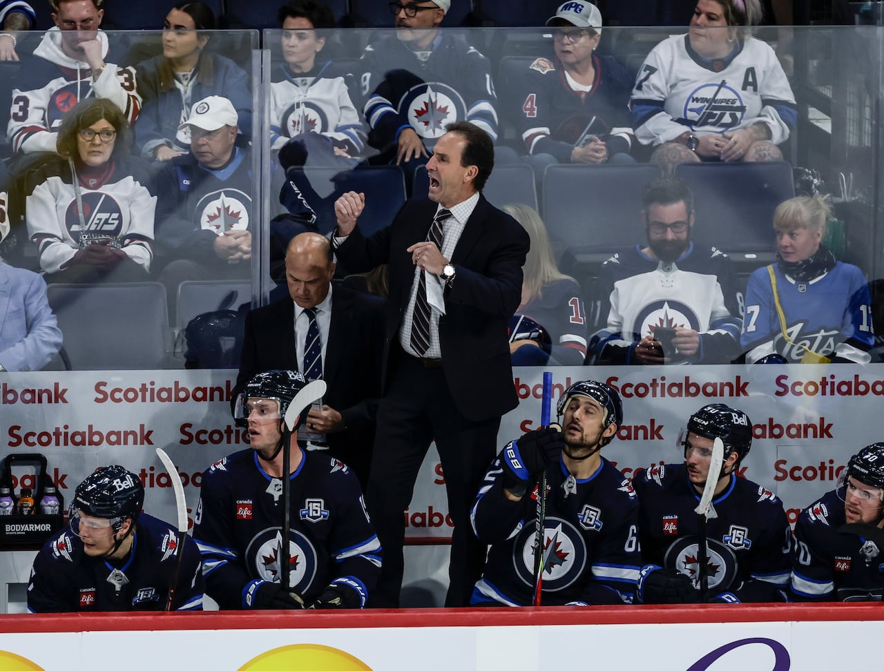 Man in a suit yells and stands on a bench lined with hockey players wearing navy blue and white jerseys
