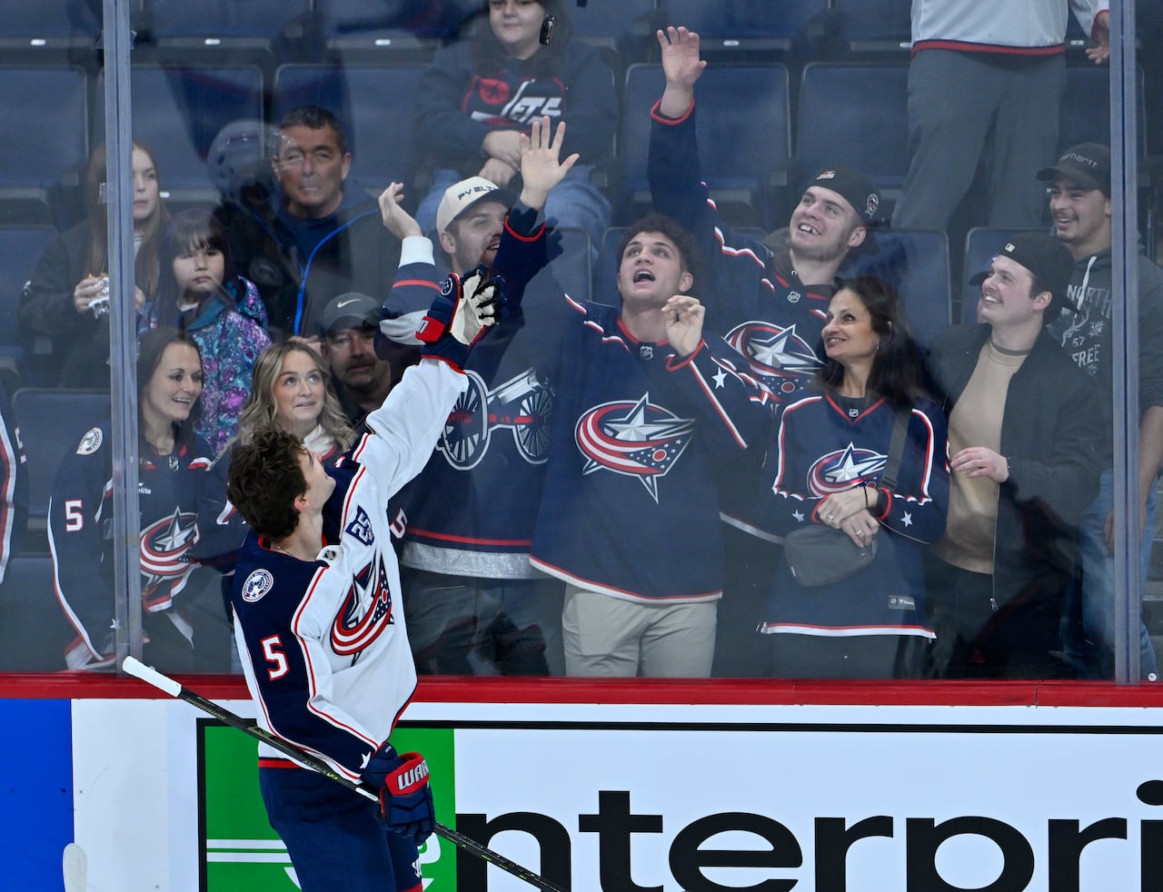 Several fans in Columbus jerseys reach up to catch a puck tossed over the glass by a helmetless Columbus player.
