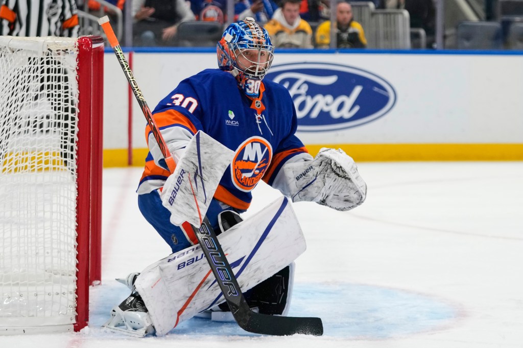A hockey goalie in blue and orange uniform and matching helmet crouches in front of the net.