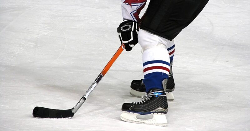 A hockey player on ice in black skates and blue-and-white socks controls a puck with a black stick, preparing to make a move during a game.