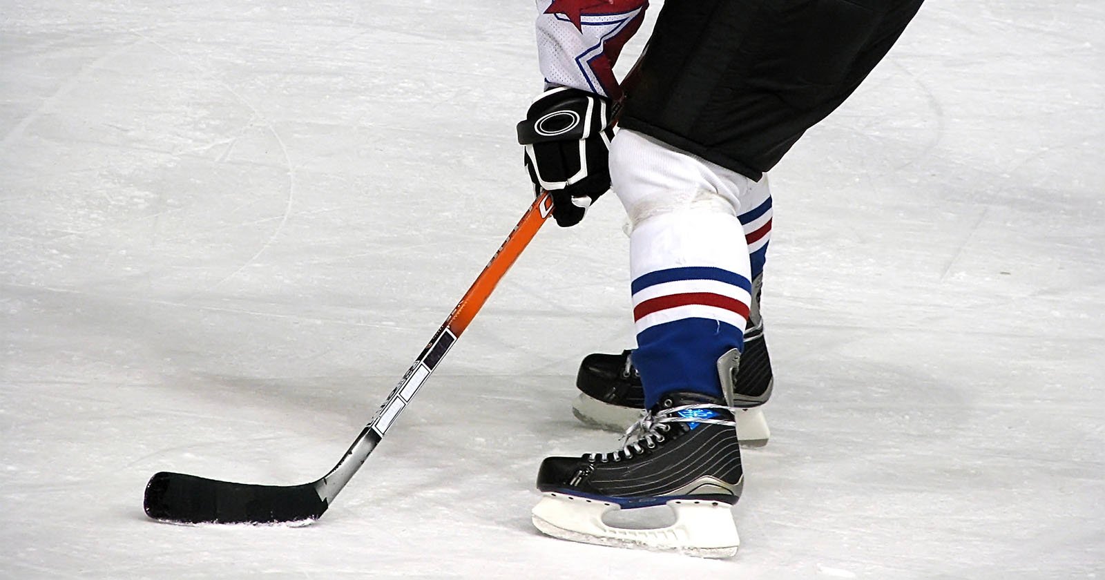 A hockey player on ice in black skates and blue-and-white socks controls a puck with a black stick, preparing to make a move during a game.
