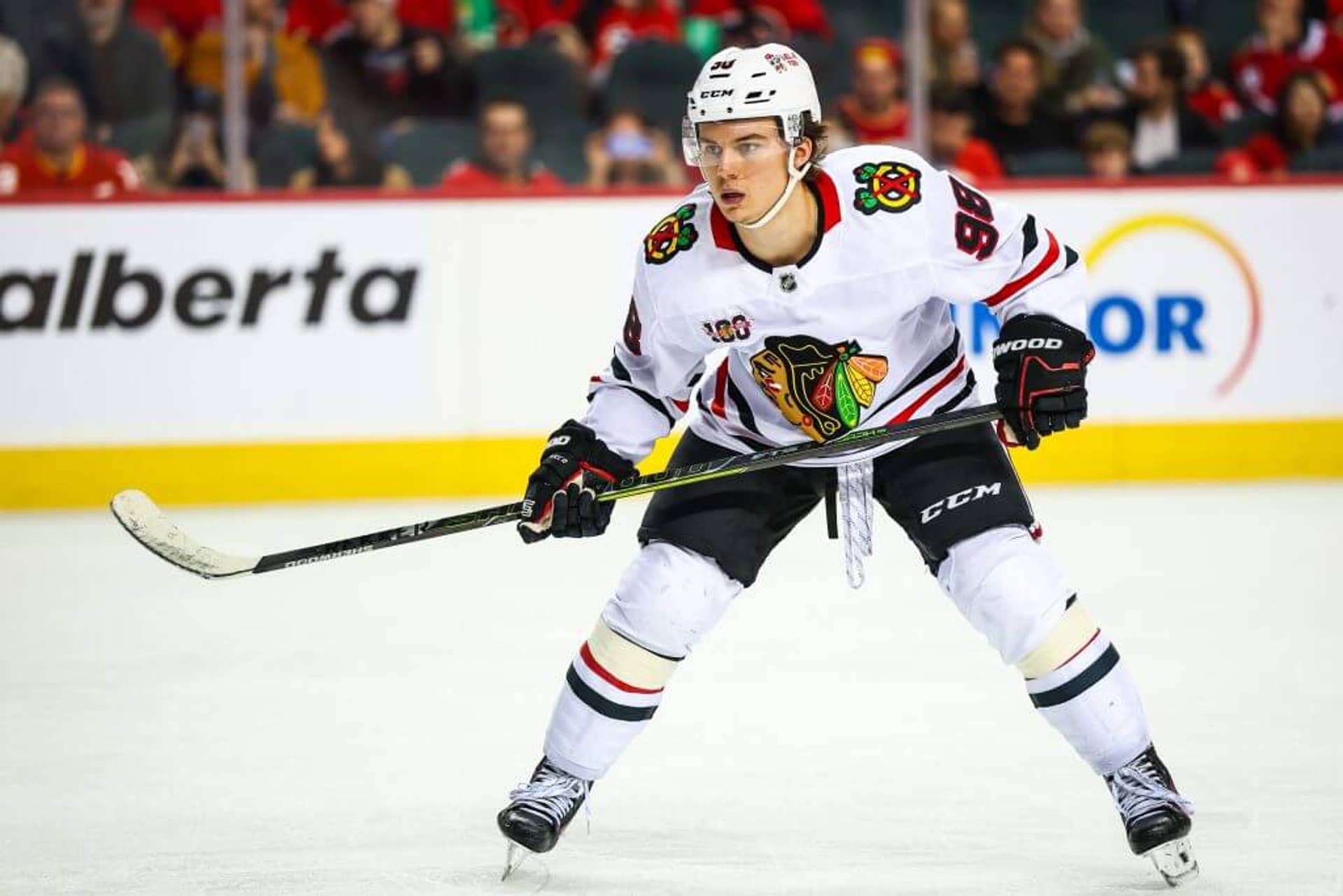Wearing a white Blackhawks jersey, Chicago forward Connor Bedard is poised center ice with his legs apart and his stick held waist high in a horizontal position.