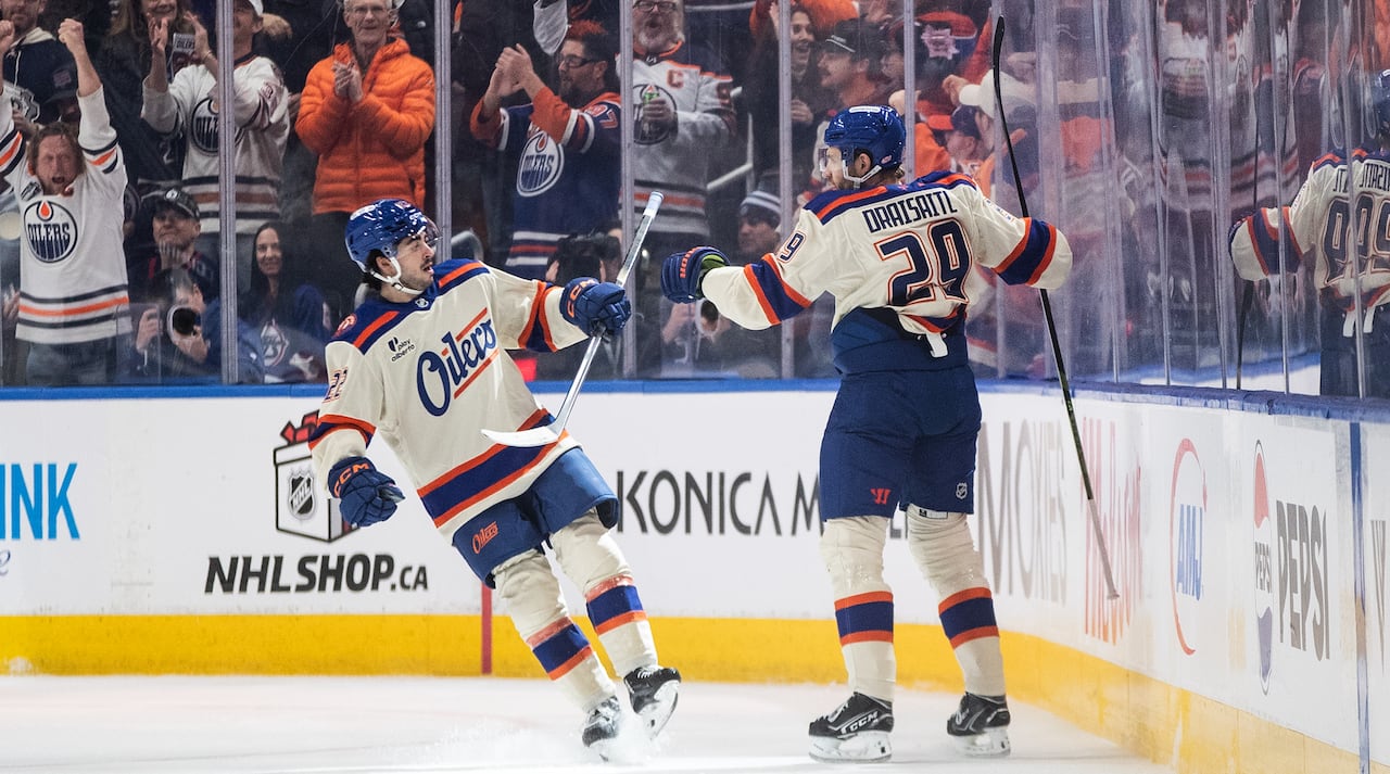 Two men in white-blue-and-orange hockey uniforms celebrate a goal on the ice. In the stands behind them, fans in team apparel are standing out of their seats, cheering.