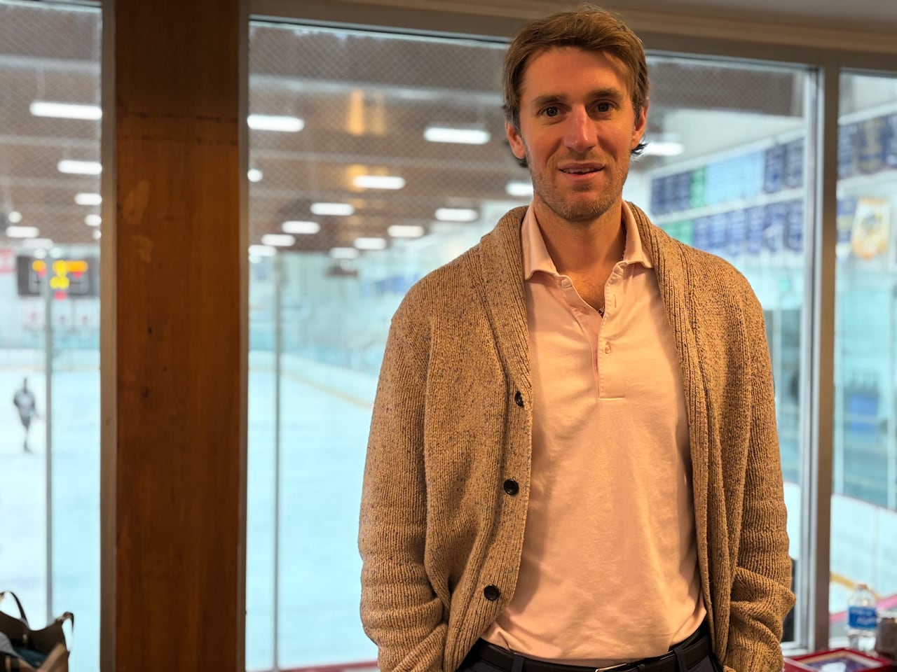 A man poses in front of a window showing an ice rink.
