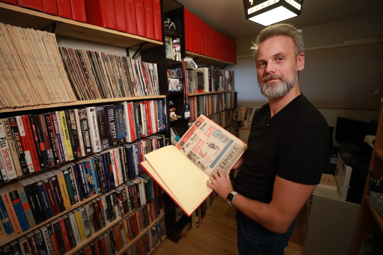 In a room full of books on bookshelves, a man in a black t-shirt and blue jeans holds open a binder with an old issue of The Hockey News inside.