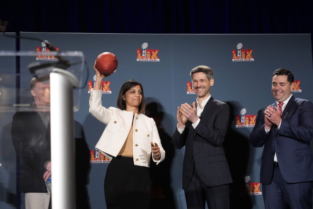 Woman wearing white jacket holding football standing on stage beside applauding men