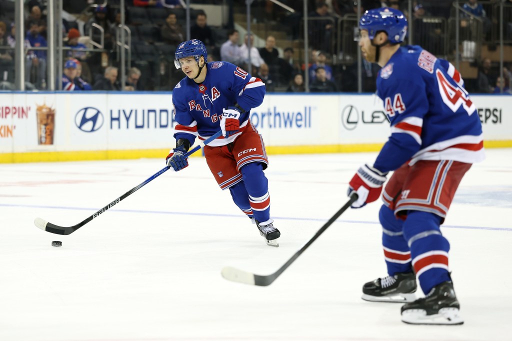 Two New York Rangers players on the ice during a game, with one player looking at the puck and holding a hockey stick.