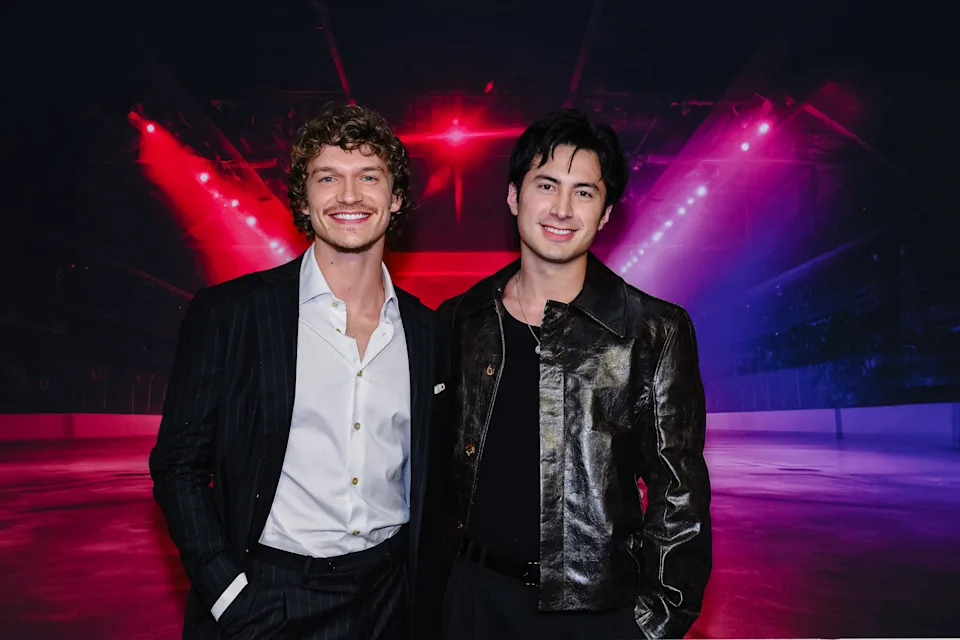 Stars Connor Storrie and Hudson Williams at the Toronto premiere of Heated Rivalry. They are at a hockey rink with bright red and purple lights behind them.