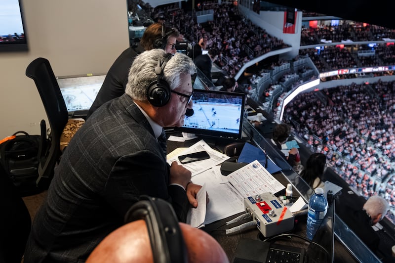 Nick Nickson from above and in profile. He is looking down from the press box at Crypto.com Arena down into a crowd of Kings fans. He wears a suit with a headset and sits at a table strewn with different papers.