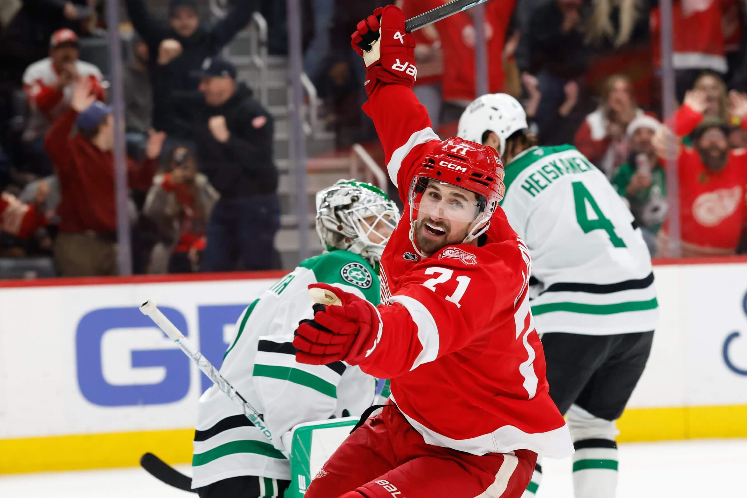 Dylan Larkin lifts his stick hand over his head in celebration with two Stars players behind him.