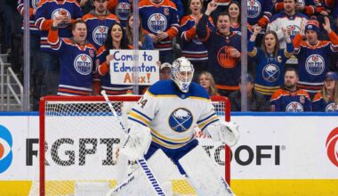 Buffalo Sabres goaltender Alex Lyon (#34), wearing his white away jersey, stands in a ready position in front of his net. Behind the protective glass, the stands are filled mostly with cheering Edmonton Oilers fans in blue and orange jerseys.