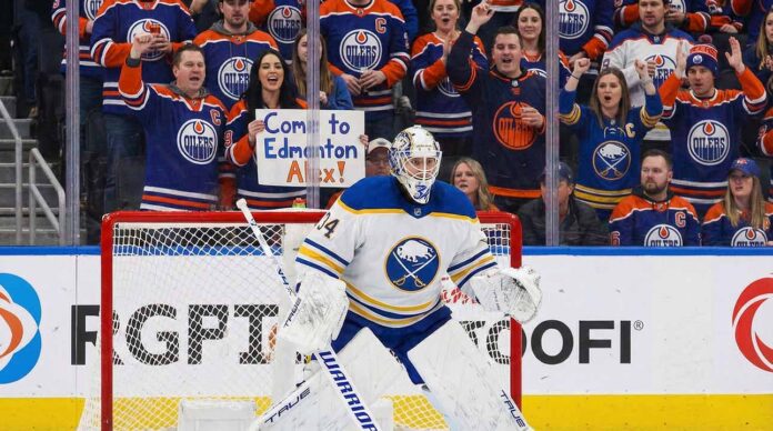 Buffalo Sabres goaltender Alex Lyon (#34), wearing his white away jersey, stands in a ready position in front of his net. Behind the protective glass, the stands are filled mostly with cheering Edmonton Oilers fans in blue and orange jerseys.