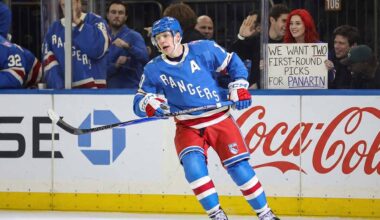 New York Rangers alternate captain Artemi Panarin (10) skating intently on the ice during a game. In the stands behind the glass, a red-haired fan holds up a white sign manually written with "WE WANT TWO FIRST-ROUND PICKS FOR PANARIN".