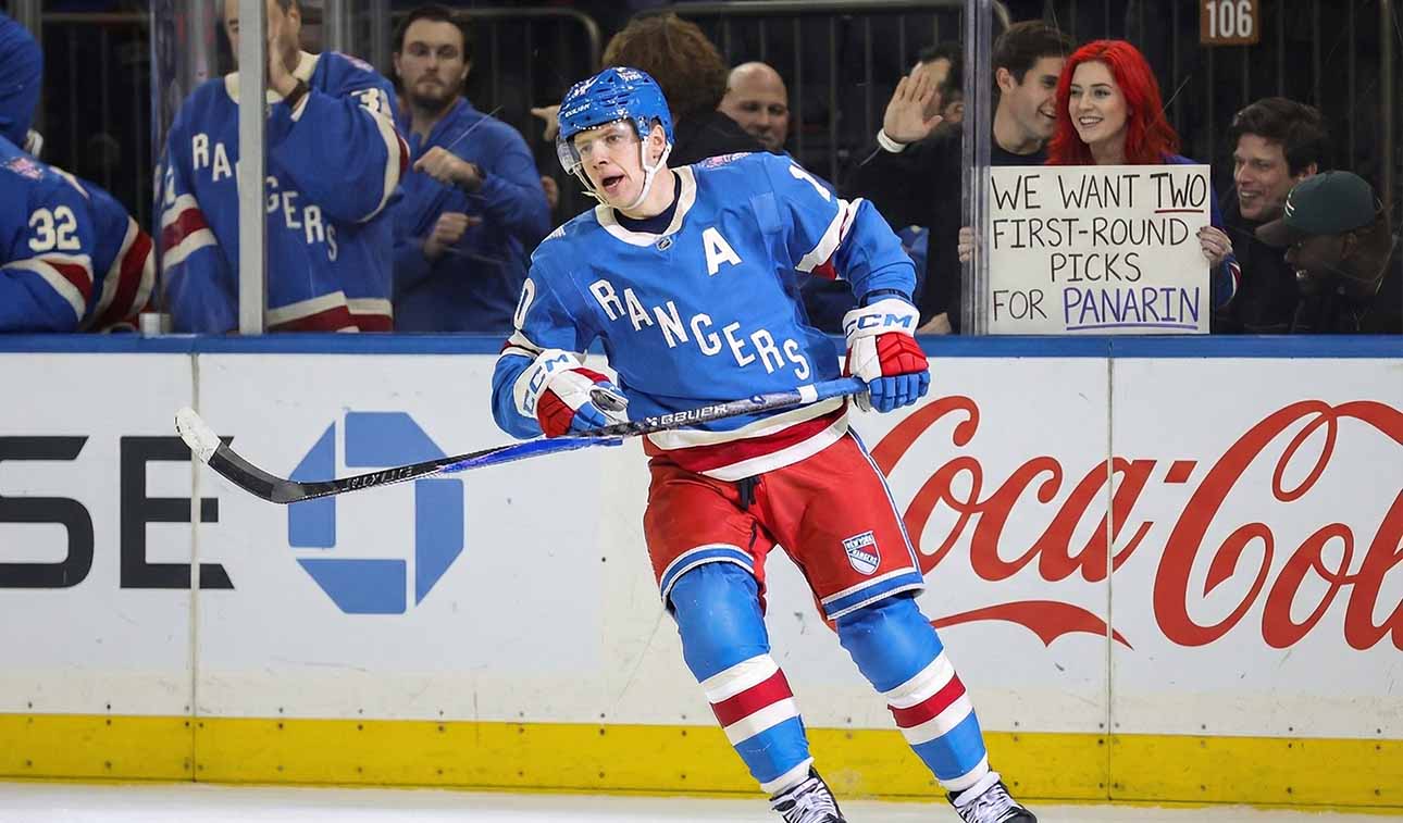 New York Rangers alternate captain Artemi Panarin (10) skating intently on the ice during a game. In the stands behind the glass, a red-haired fan holds up a white sign manually written with "WE WANT TWO FIRST-ROUND PICKS FOR PANARIN".