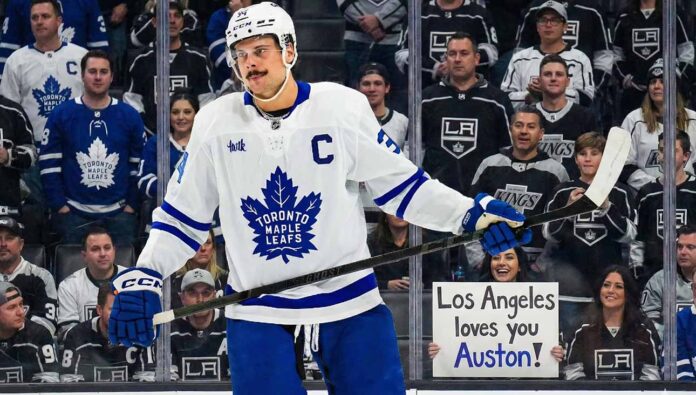 Toronto Maple Leafs captain Auston Matthews stands on the ice in his white away jersey, looking towards the stands behind the glass. The crowd features numerous Los Angeles Kings fans, and a smiling woman in the front row holds up a handwritten sign that reads, "Los Angeles loves you Auston!", amidst ongoing trade speculation.