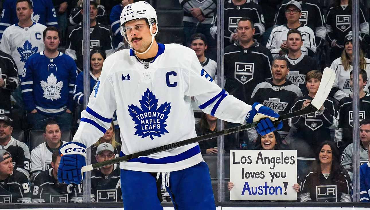 Toronto Maple Leafs captain Auston Matthews stands on the ice in his white away jersey, looking towards the stands behind the glass. The crowd features numerous Los Angeles Kings fans, and a smiling woman in the front row holds up a handwritten sign that reads, "Los Angeles loves you Auston!", amidst ongoing trade speculation.