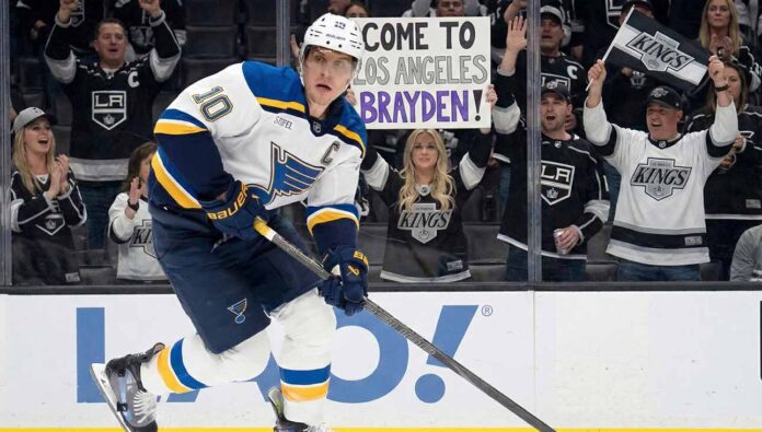 St. Louis Blues captain Brayden Schenn (#10) skates with the puck past tall rink boards featuring a Coca-Cola advertisement. Behind the glass, Los Angeles Kings fans cheer, and a woman holds up a handmade sign that reads "COME TO LOS ANGELES BRAYDEN!".