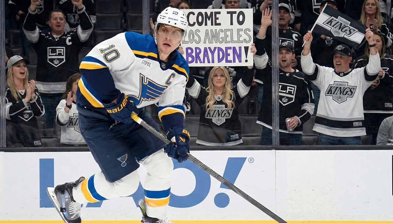 St. Louis Blues captain Brayden Schenn (#10) skates with the puck past tall rink boards featuring a Coca-Cola advertisement. Behind the glass, Los Angeles Kings fans cheer, and a woman holds up a handmade sign that reads "COME TO LOS ANGELES BRAYDEN!".