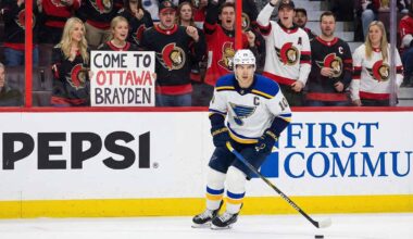 St. Louis Blues forward Brayden Schenn, wearing his white away jersey, skates with the puck during a game. In the stands behind the glass, a crowd of Ottawa Senators fans cheer, and a blonde woman in the front row holds up a white sign with red and black text that reads, "COME TO OTTAWA BRAYDEN".
