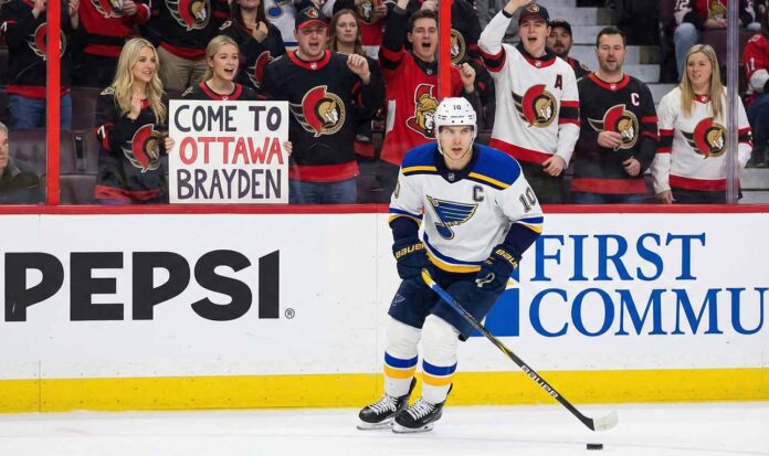 St. Louis Blues forward Brayden Schenn, wearing his white away jersey, skates with the puck during a game. In the stands behind the glass, a crowd of Ottawa Senators fans cheer, and a blonde woman in the front row holds up a white sign with red and black text that reads, "COME TO OTTAWA BRAYDEN".
