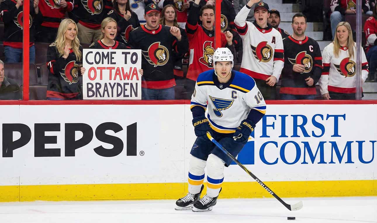 St. Louis Blues forward Brayden Schenn, wearing his white away jersey, skates with the puck during a game. In the stands behind the glass, a crowd of Ottawa Senators fans cheer, and a blonde woman in the front row holds up a white sign with red and black text that reads, "COME TO OTTAWA BRAYDEN".