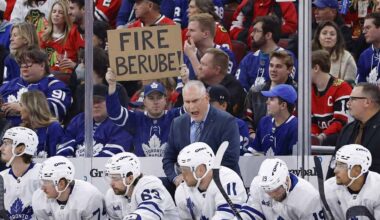 Toronto Maple Leafs head coach Craig Berube yells angrily from behind the players' bench during a game. Directly behind him in the stands, a fan holds up a handwritten cardboard sign that explicitly reads "FIRE BERUBE!".