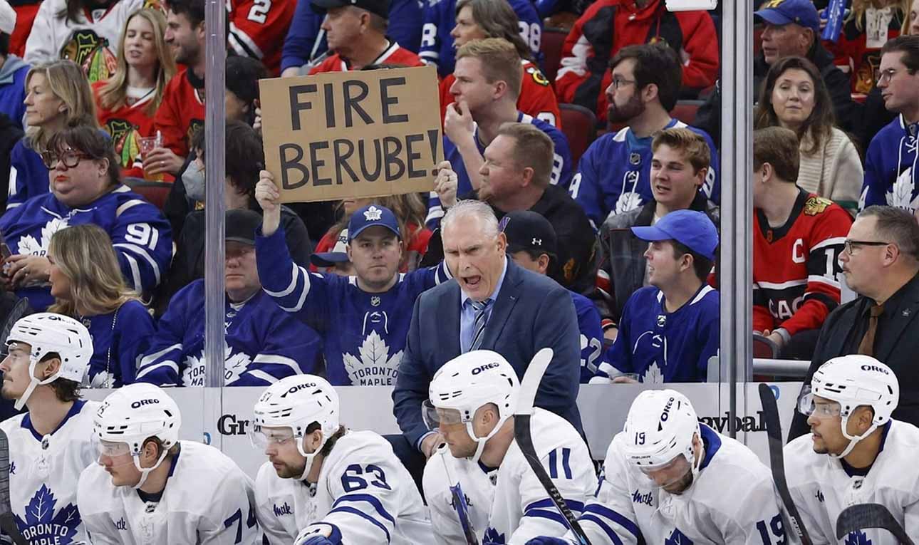 Toronto Maple Leafs head coach Craig Berube yells angrily from behind the players' bench during a game. Directly behind him in the stands, a fan holds up a handwritten cardboard sign that explicitly reads "FIRE BERUBE!".