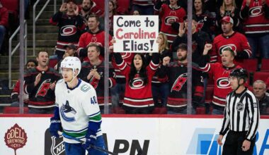 Vancouver Canucks forward Elias Pettersson (40) skates on the ice during a game. Behind the glass, a female fan in a Carolina Hurricanes jersey holds up a sign that reads, "Come to Carolina Elias!", while surrounded by other cheering Hurricanes fans.