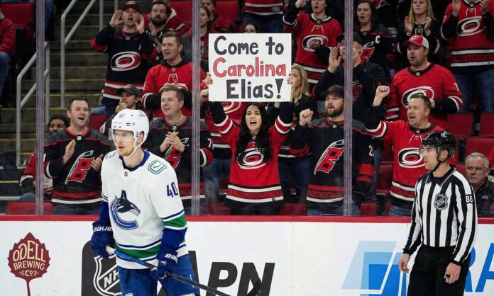 Vancouver Canucks forward Elias Pettersson (40) skates on the ice during a game. Behind the glass, a female fan in a Carolina Hurricanes jersey holds up a sign that reads, "Come to Carolina Elias!", while surrounded by other cheering Hurricanes fans.