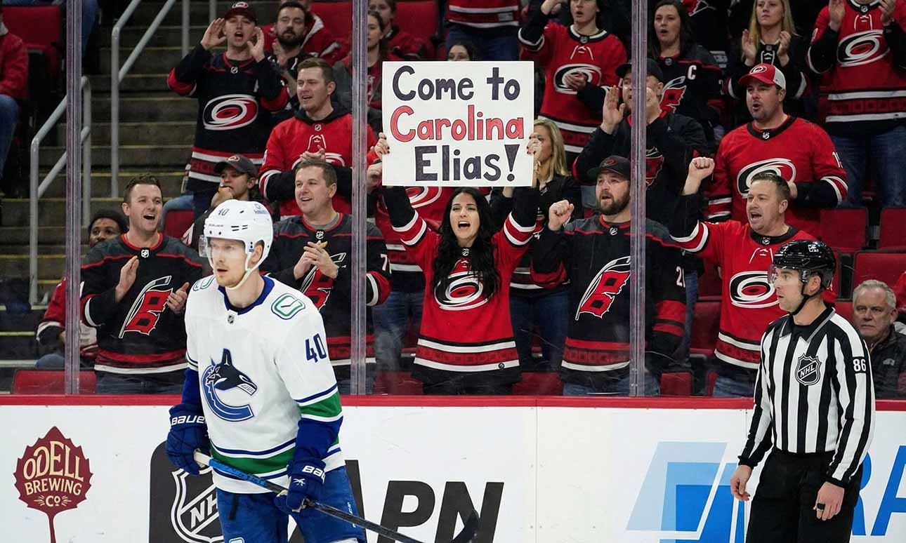 Vancouver Canucks forward Elias Pettersson (40) skates on the ice during a game. Behind the glass, a female fan in a Carolina Hurricanes jersey holds up a sign that reads, "Come to Carolina Elias!", while surrounded by other cheering Hurricanes fans.