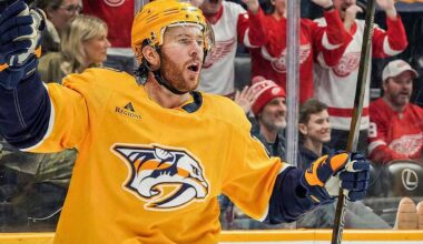 Jonathan Marchessault of the Nashville Predators, wearing a yellow jersey, celebrates on the ice with his mouth open and stick raised. Behind the glass, fans wearing red Detroit Red Wings apparel cheer.