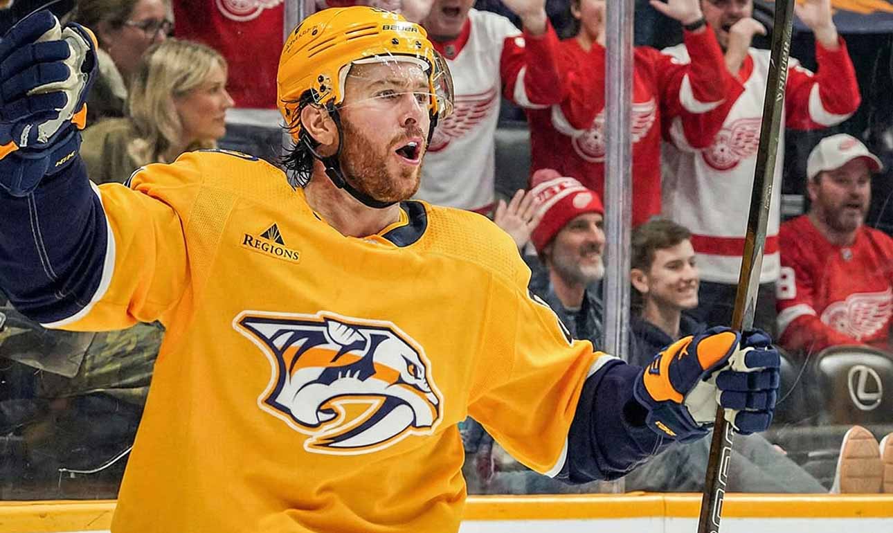 Jonathan Marchessault of the Nashville Predators, wearing a yellow jersey, celebrates on the ice with his mouth open and stick raised. Behind the glass, fans wearing red Detroit Red Wings apparel cheer.