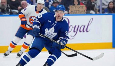 Toronto Maple Leafs Mattias Maccelli in a blue jersey, number 63, skates with the puck during a hockey game. Behind him, a New York Islanders player in a white jersey, number 27, follows. In the background stands, a fan holds up a cardboard sign that reads "BYE-BYE MATTIAS!".