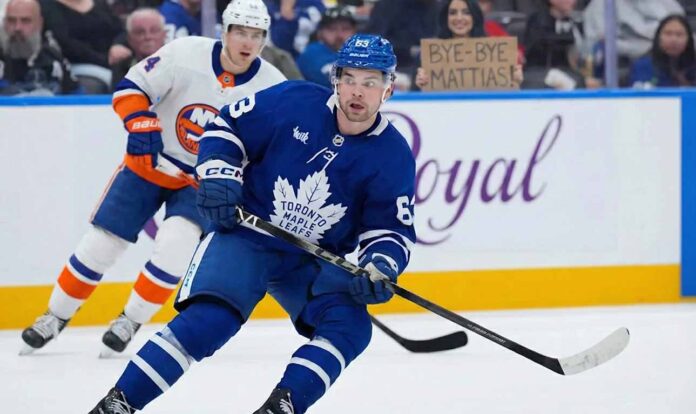 Toronto Maple Leafs Mattias Maccelli in a blue jersey, number 63, skates with the puck during a hockey game. Behind him, a New York Islanders player in a white jersey, number 27, follows. In the background stands, a fan holds up a cardboard sign that reads "BYE-BYE MATTIAS!".