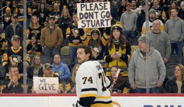 Stuart Skinner, wearing his white #74 Pittsburgh Penguins jersey, stands on the ice looking towards the fans behind the plexiglass. A fan in the center holds up a white sign with black letters that reads "PLEASE DON'T GO STUART".