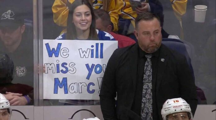ormer Toronto Maple Leafs assistant coach Marc Savard stands on the bench looking serious while a fan behind the glass holds a handwritten sign reading We will miss you Marc.