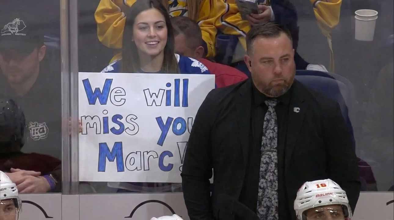 ormer Toronto Maple Leafs assistant coach Marc Savard stands on the bench looking serious while a fan behind the glass holds a handwritten sign reading We will miss you Marc.