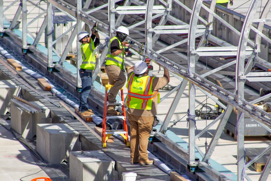Three men in yellow and orange safety vests and white hardhats use tools to build the metal frame structure of a massive tent. 