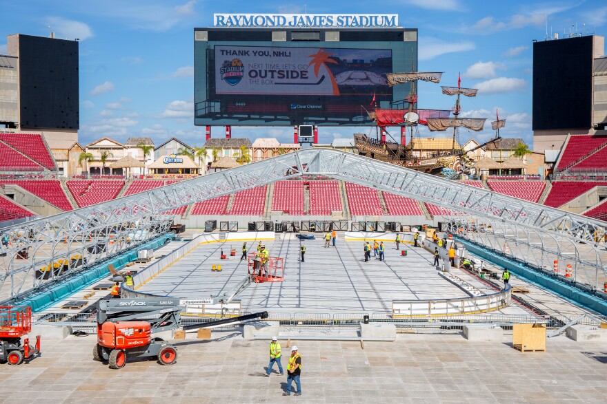 A football field stripped bare of its turf and several tiers of red stadium seating in the background. On the ground floor, the metal skeleton of what will be a climate-controlled tent is partially built. 