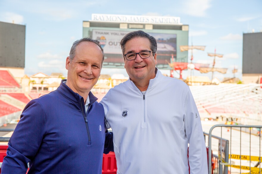 Two smiling middle age white men stand side by side with a looming football stadium in the background. 
