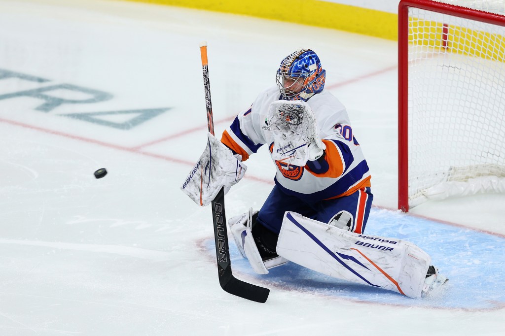 A New York Islanders goalie in a white and blue uniform makes a save, deflecting the puck with their stick.