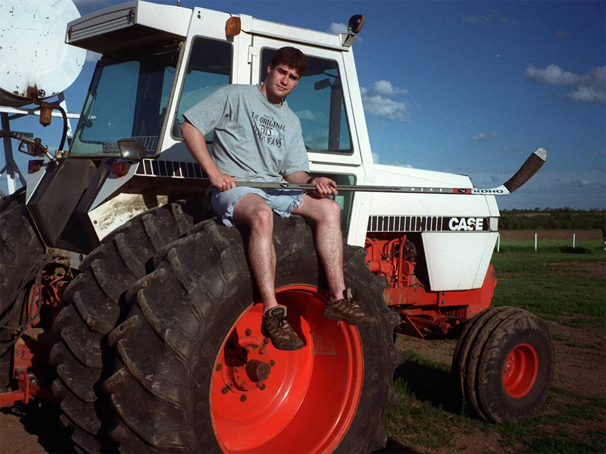 A young Patrick Marleau sits on the dual rear wheels of a Case tractor in shorts and a t-shirt holding a hockey stick.