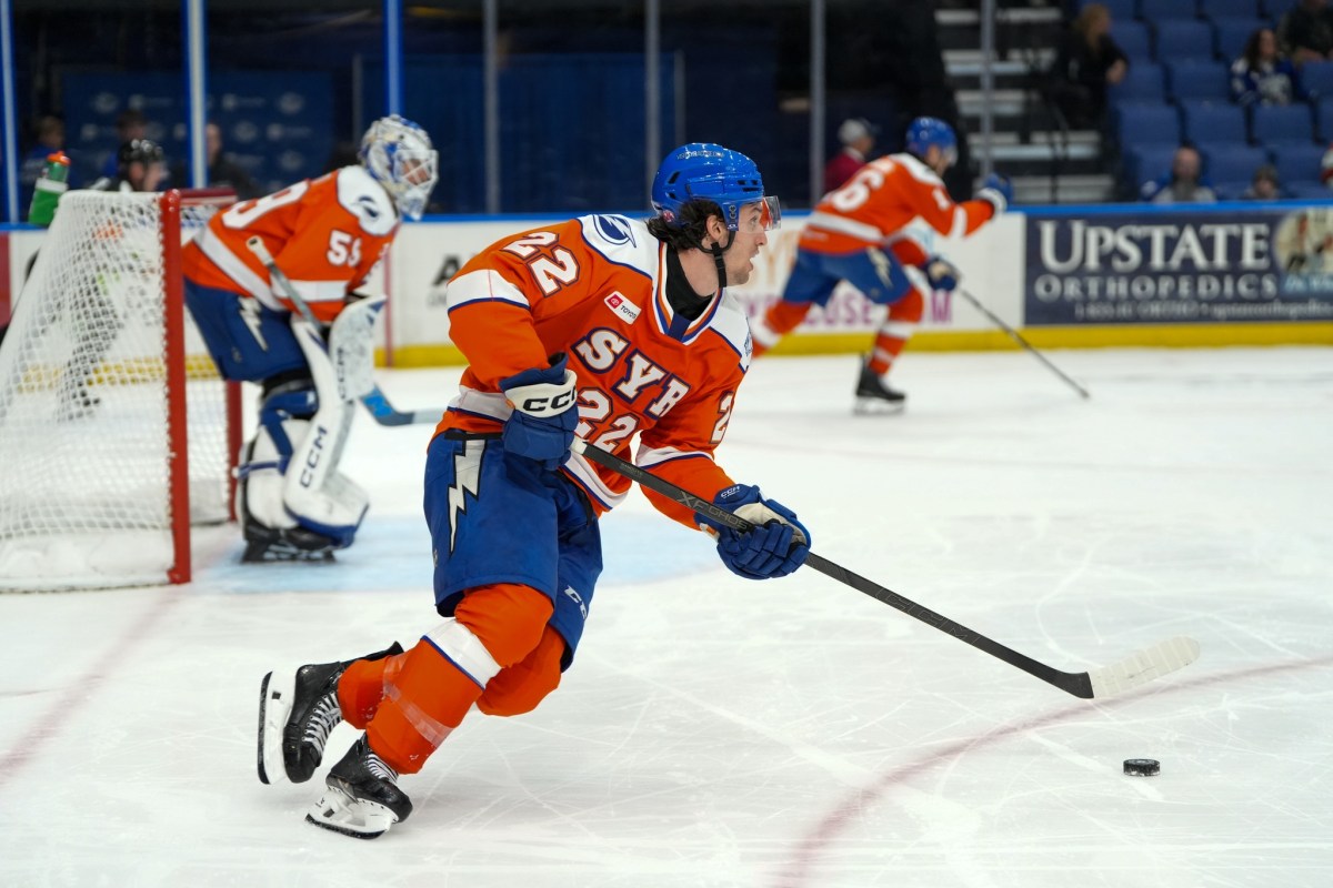 A hockey player in an orange and blue uniform skates up the ice with the puck in front of him