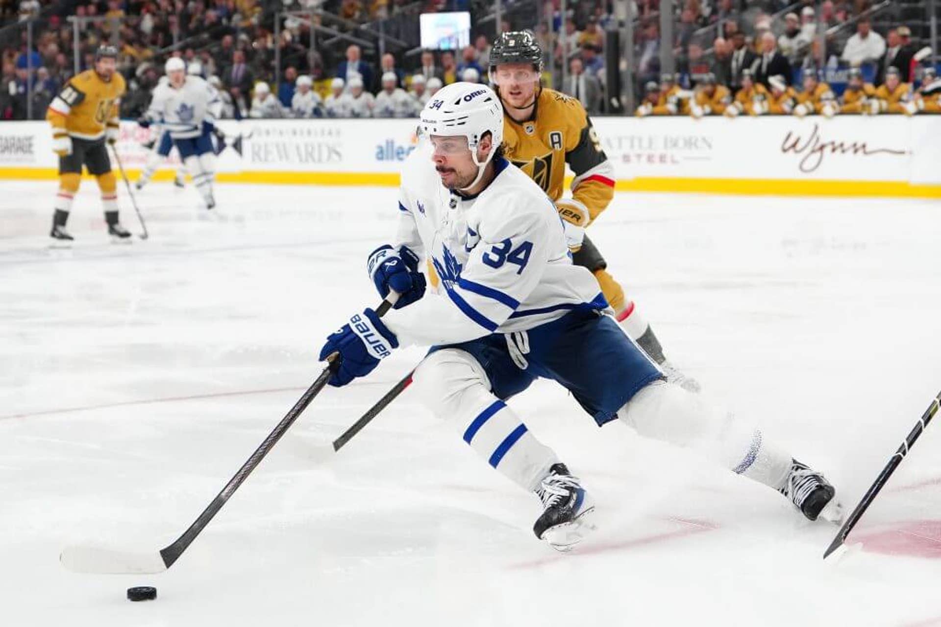 Toronto center Auston Matthews, donning a white Maple Leafs jersey and helmet with blue pants, starts to make a cut, his stick crossed over his body to his right side as he handles the puck.