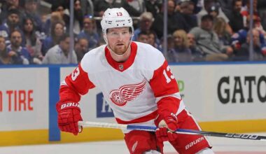Detroit Red Wings forward Alexis Lafrenière, wearing number 13 in a white away jersey with red trim and the winged wheel logo, skates on the ice holding his stick during a game.